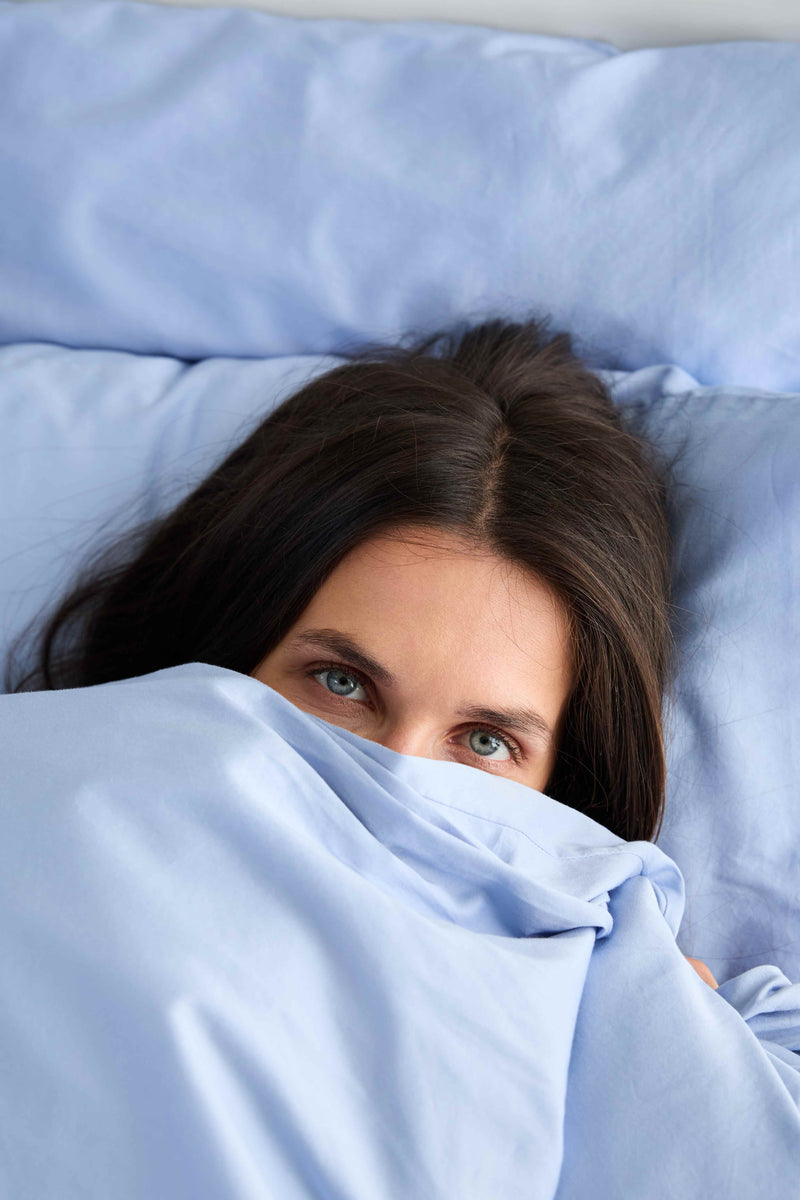 Close-up portrait of a woman peeking over light blue organic cotton sateen bedding