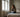 Bright European bedroom with a woman sitting on the bed rubbing her nose, faint mould spots near the window and soft neutral bedding.
