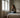 Bright European bedroom with a woman sitting on the bed rubbing her nose, faint mould spots near the window and soft neutral bedding.